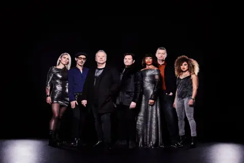 I membri dei Simple Minds posano in gruppo su sfondo nero, vestiti con abiti eleganti e scintillanti.