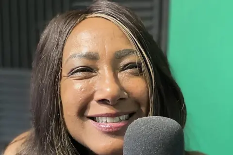 Dee C. Lee sorride vicino a un microfono, con uno sfondo verde e pannelli acustici.