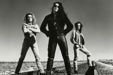 I membri dei The Cult posano sulle rotaie in un paesaggio desertico, con un cielo limpido sullo sfondo.