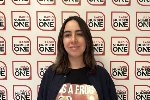 Federica Abbate, con una maglietta a tema rana, sorride davanti a uno sfondo con il logo di Radio Number One. Le sue canzoni risuonano in radio.