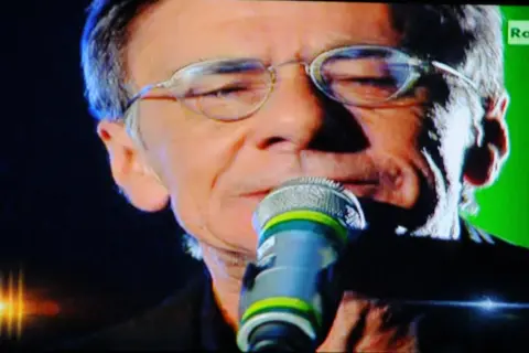 Roberto Vecchioni canta "Chiamami ancora amore" con passione, il microfono vicino alle labbra, sullo sfondo verde di Rai 1.