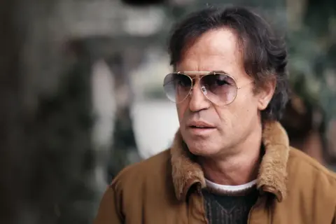 Close-up of a man with dark hair, wearing aviator sunglasses and a brown jacket with a fur collar.