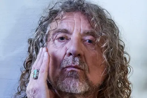 Primo piano di Robert Plant, con la mano sul viso, un anello turchese. La sua espressione è intensa, quasi una saving grace per chi lo osserva.