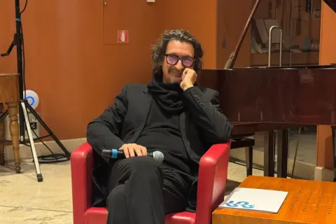 Sergio Cammariere, con microfono in mano, siede su una poltrona rossa, pronto a raccontare le sue canzoni.
