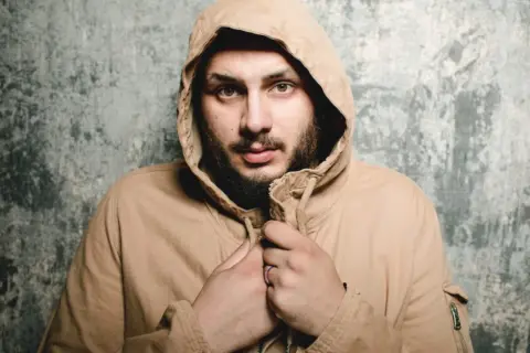 Uomo con cappuccio beige, sguardo intenso, barba. Sembra che qualcuno abbia sbagliato con lui.