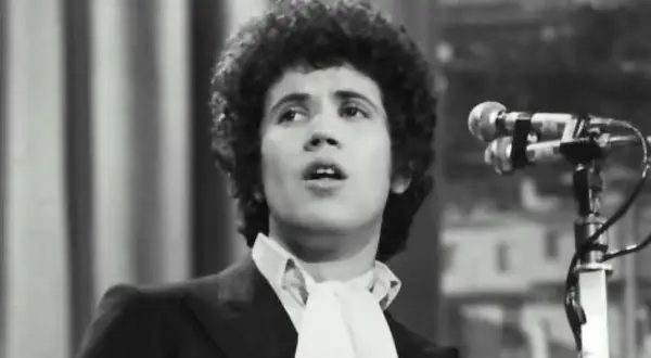 Lucio Battisti, con la sua iconica chioma, canta "Il tempo di morire" davanti a un microfono.