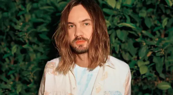 Kevin Parker, uno dei membri dei Tame Impala, con lunghi capelli castani e una camicia floreale, sullo sfondo di un muro di foglie verdi.