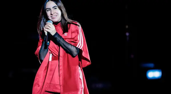 Federica Abbate canta sul palco, sorridente, con un outfit rosso Adidas. I suoi amici la guardano con ammirazione.