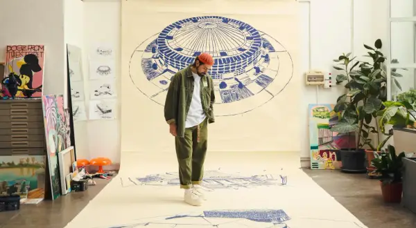 Un artista con un cappello arancione osserva disegni architettonici su carta, come se stesse creando nuove canzoni di Frah Quintale.