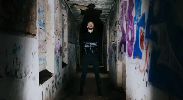 Uomo con barba in piedi in un corridoio pieno di graffiti, con indosso un abito nero. Sembra un rapper pronto per una sfida.