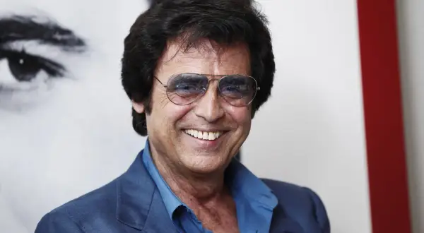 Un uomo sorridente con occhiali da sole aviator, indossa una giacca blu. Potrebbe essere Little Tony, pronto a cantare le sue canzoni.