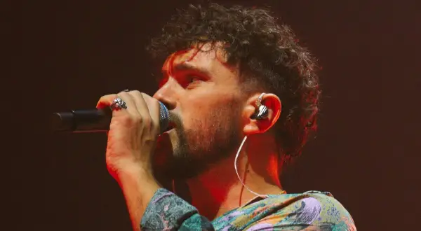 Marco Castello canta con passione, microfono in mano, cuffie nell'orecchio e un look colorato.
