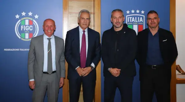 Quattro uomini in posa davanti a uno sfondo blu con il logo FIGC. Nessuno sembra un "dead can dance", ma tutti sono vestiti elegantemente.