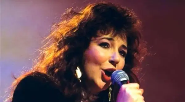 Kate Bush ora, con i suoi capelli scuri e fluenti, canta con passione al microfono, illuminata da luci calde.