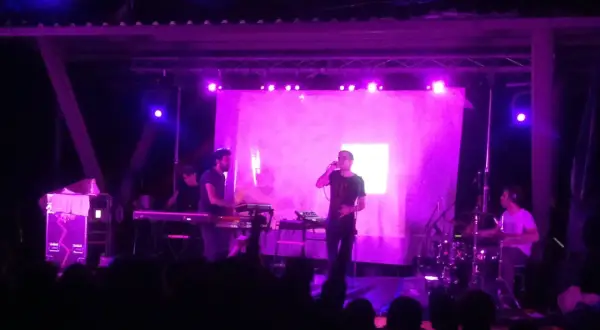 Concerto con luci viola. Sul palco, un cantante con occhiali e una tastiera. Dietro, un batterista e altri musicisti. Il pubblico è in ombra.