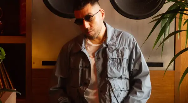 Man in sunglasses and a grey jacket leans against a large speaker.