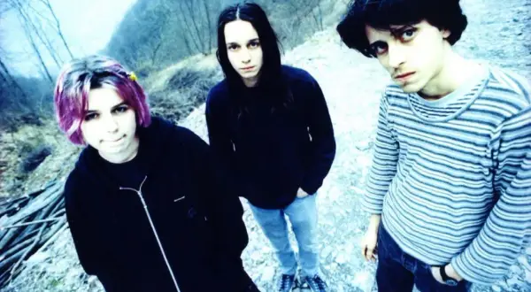 Three young people with dark hair and casual clothing stand outdoors in a rocky, wooded area.