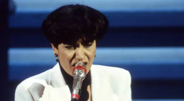 Mia Martini, icona della musica italiana, canta con passione sul palco, illuminata da luci soffuse.