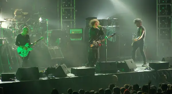 Sul palco, i membri dei The Cure suonano le loro iconiche canzoni. Un chitarrista con una chitarra verde brillante, il cantante con la sua chitarra nera e un altro chitarrista.