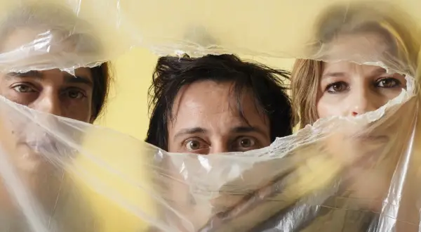Occhi curiosi di Alberto Ferrari Verdena e altri due volti emergono da un telo di plastica trasparente su sfondo giallo.