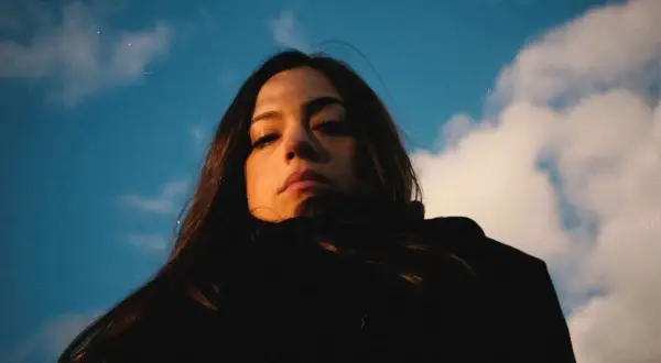 Daniela Pes spira sotto un cielo azzurro e nuvoloso, con uno sguardo intenso e capelli scuri che incorniciano il viso.