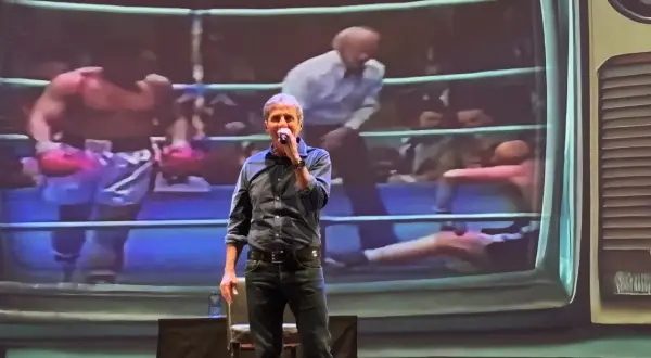 Uomo sul palco con microfono, sullo sfondo un ring di pugilato proiettato. "Dai mamma dai", urla la folla.