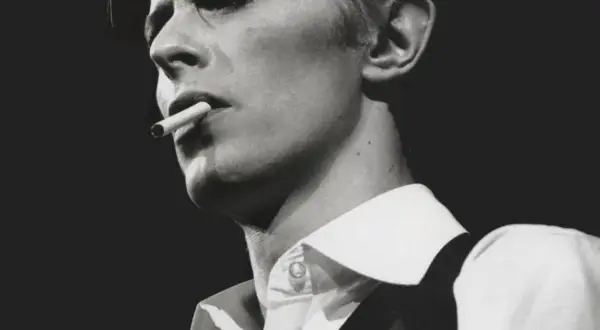 David Bowie, un'icona dei suoi golden years, produttore di musica e stile, fuma una sigaretta con sguardo intenso.