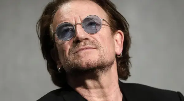 Primo piano di Bono Vox con occhiali rotondi blu, che ricorda le sue iconiche canzoni.