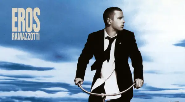 Copertina album di Eros Ramazzotti con lui in giacca e cravatta su una bicicletta sotto un cielo blu.