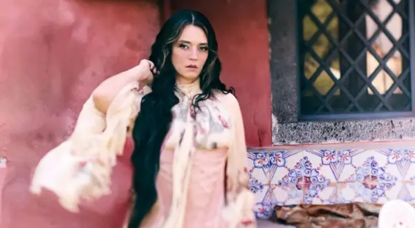 Carmen Consoli, con lunghi capelli scuri e un abito etereo, posa con un'espressione intensa. L'ambientazione ricorda un cortile con piastrelle decorative, evocando un'atmosfera da "Orfeo".
