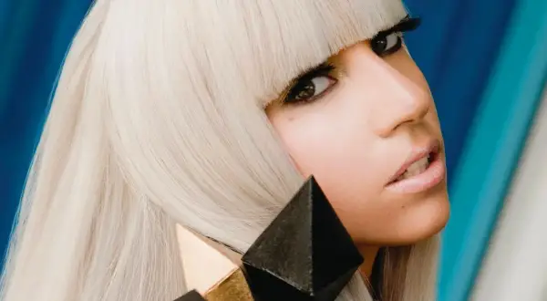 Lady Gaga, con capelli biondo platino e un look audace, cattura l'essenza del suo **inizio carriera** con un'espressione intensa e un accessorio geometrico.