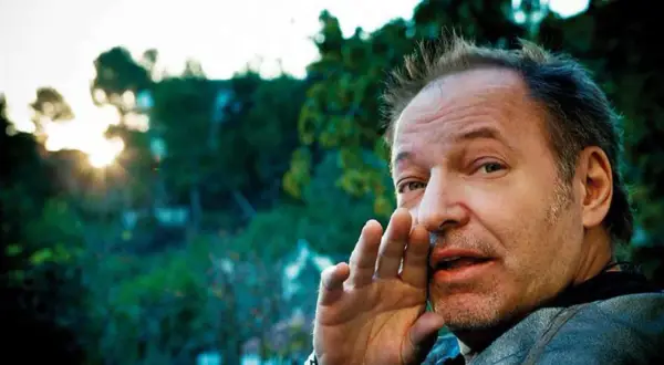Vasco Rossi sussurra "ti prendo e ti porto via" con la mano all'orecchio, mentre il sole tramonta tra gli alberi.