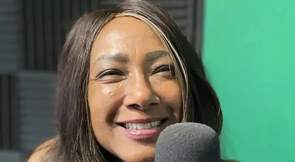 Dee C. Lee sorride vicino a un microfono, con uno sfondo verde e pannelli acustici.