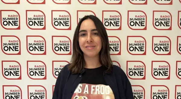 Federica Abbate, con una maglietta a tema rana, sorride davanti a uno sfondo con il logo di Radio Number One. Le sue canzoni risuonano in radio.