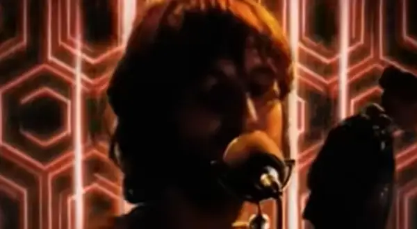 Un uomo con capelli castani canta al microfono, sullo sfondo un motivo esagonale rosso. Sembra che baustelle charlie fa surf con la sua musica.