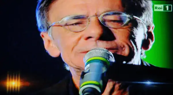 Roberto Vecchioni canta "Chiamami ancora amore" con passione, il microfono vicino alle labbra, sullo sfondo verde di Rai 1.