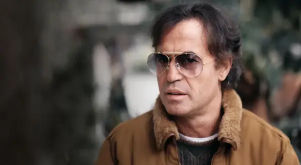 Close-up of a man with dark hair, wearing aviator sunglasses and a brown jacket with a fur collar.