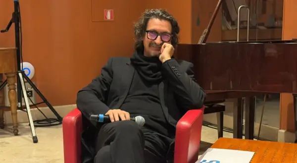 Sergio Cammariere, con microfono in mano, siede su una poltrona rossa, pronto a raccontare le sue canzoni.