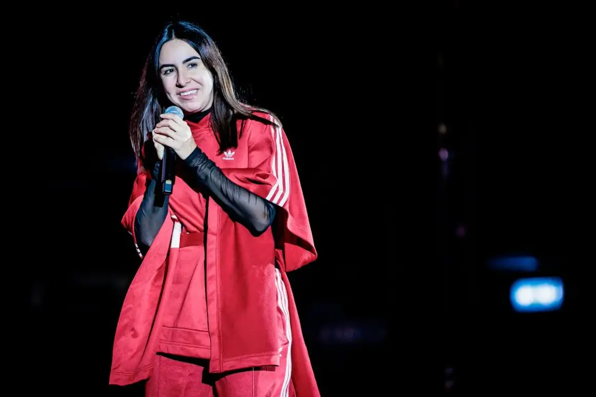Federica Abbate canta sul palco, sorridente, con un outfit rosso Adidas. I suoi amici la guardano con ammirazione.