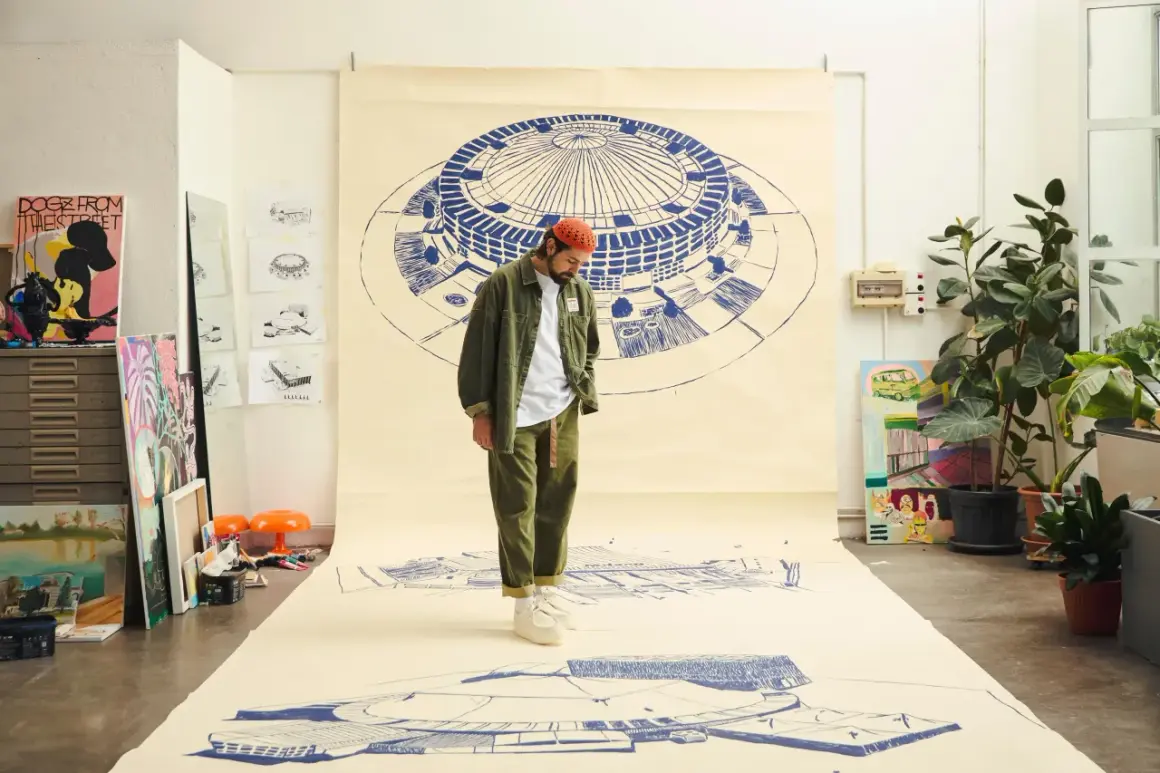 Un artista con un cappello arancione osserva disegni architettonici su carta, come se stesse creando nuove canzoni di Frah Quintale.