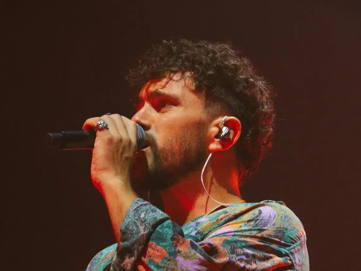 Marco Castello canta con passione, microfono in mano, cuffie nell'orecchio e un look colorato.