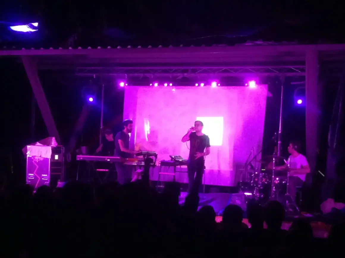 Concerto con luci viola. Sul palco, un cantante con occhiali e una tastiera. Dietro, un batterista e altri musicisti. Il pubblico è in ombra.
