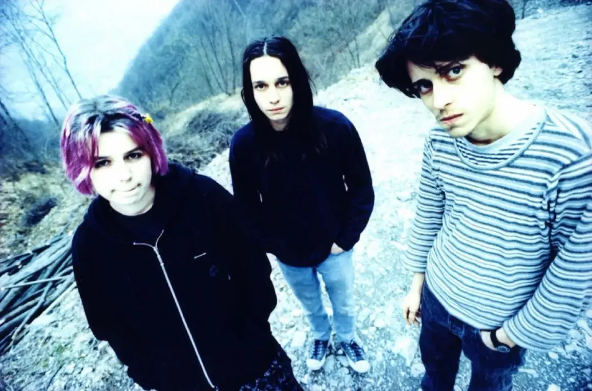 Three young people with dark hair and casual clothing stand outdoors in a rocky, wooded area.