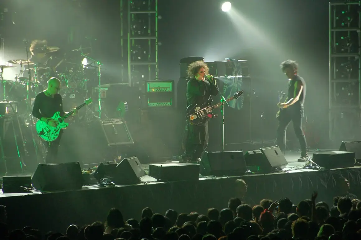 Sul palco, i membri dei The Cure suonano le loro iconiche canzoni. Un chitarrista con una chitarra verde brillante, il cantante con la sua chitarra nera e un altro chitarrista.