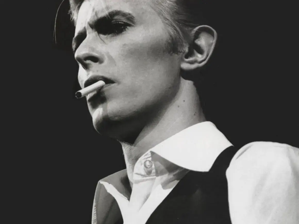 David Bowie, un'icona dei suoi golden years, produttore di musica e stile, fuma una sigaretta con sguardo intenso.