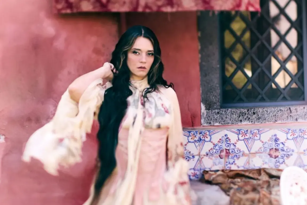 Carmen Consoli, con lunghi capelli scuri e un abito etereo, posa con un'espressione intensa. L'ambientazione ricorda un cortile con piastrelle decorative, evocando un'atmosfera da "Orfeo".