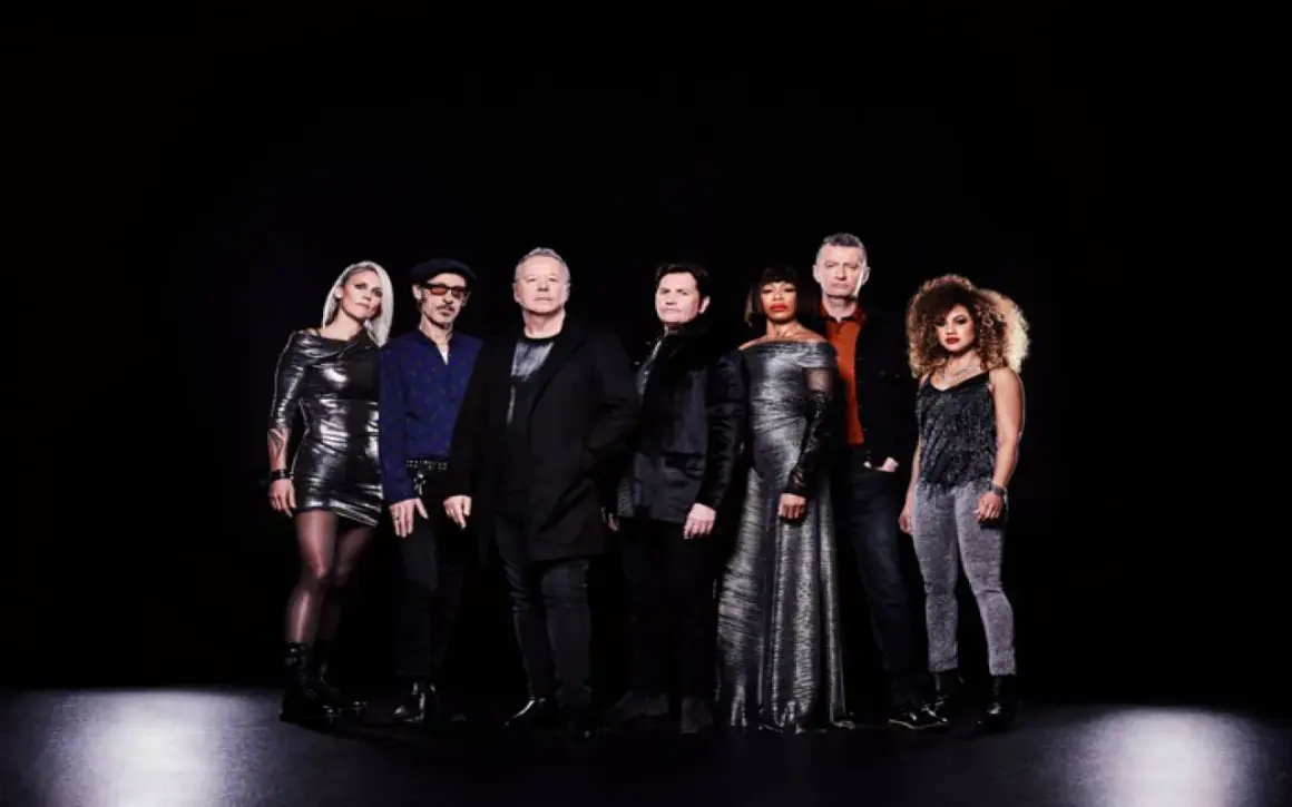 I membri dei Simple Minds posano in gruppo su sfondo nero, vestiti con abiti eleganti e scintillanti.