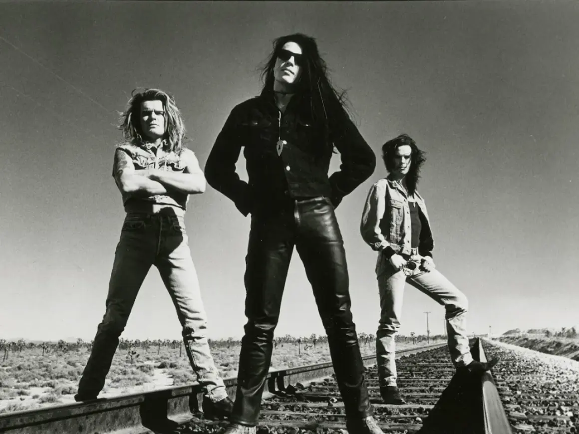 I membri dei The Cult posano sulle rotaie in un paesaggio desertico, con un cielo limpido sullo sfondo.