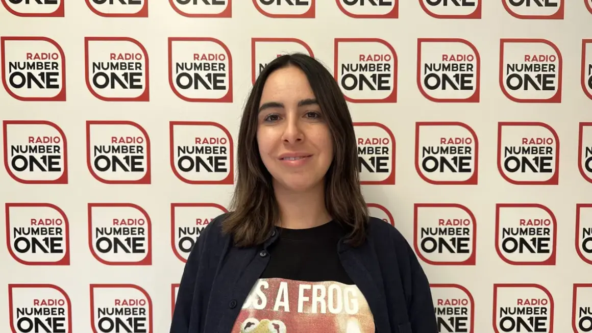Federica Abbate, con una maglietta a tema rana, sorride davanti a uno sfondo con il logo di Radio Number One. Le sue canzoni risuonano in radio.