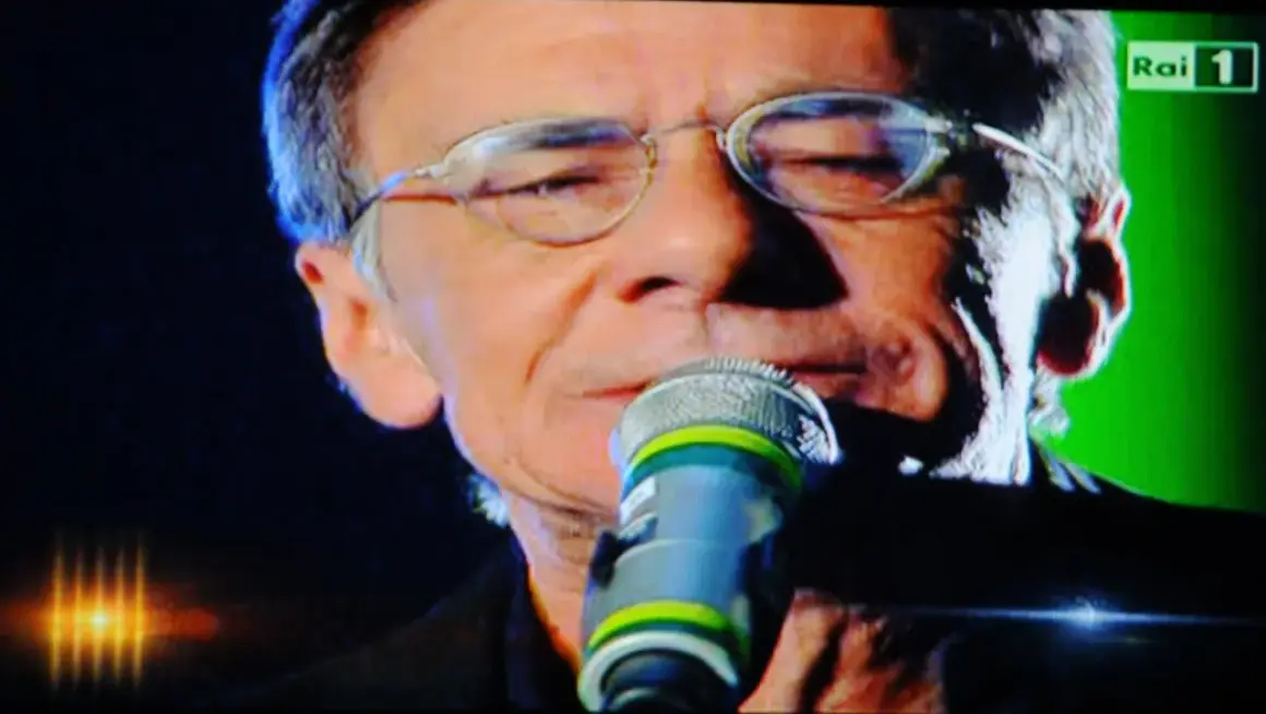 Roberto Vecchioni canta "Chiamami ancora amore" con passione, il microfono vicino alle labbra, sullo sfondo verde di Rai 1.